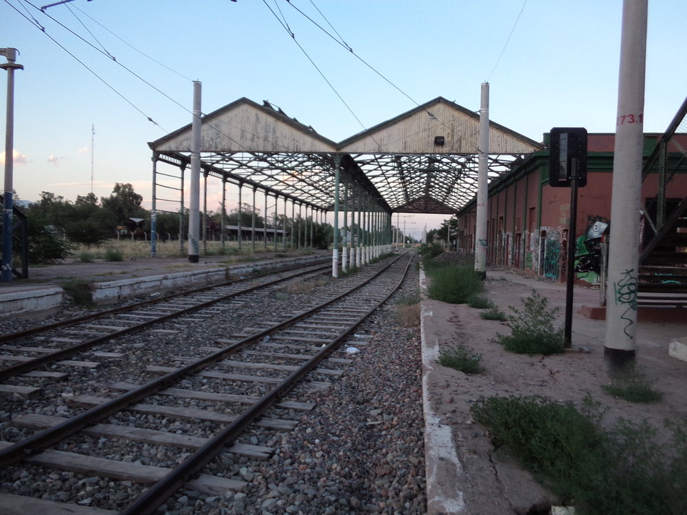 Průjezdné nádraží Mendoza patřilo dráze zvané Ferrocarril General San Martín (rozchod 1 676 mm). Končily zde mimo jiné i vlaky z Buenos Aires, ze kterého se sem dalo dostat z obou možných směrů, neboť Mendozu obklopoval jakýsi železniční okruh, jak už ostatně vyplynulo z jednoho z předchozích komentářů. Zaústěna sem byla i větev dráhy Ferrocarril General Belgrano (rozchod 1 000 mm), byť hlavní stanice této dráhy byla v Mendoze (přesněji řečeno na území Gran Mendozy) někde jinde, a sice asi 3 km východně od této (tj. napravo od snímku).