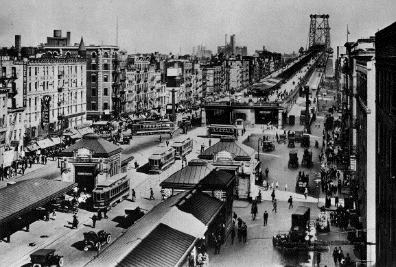 Dopravní ruch u mostu Williamsburg Bridge v roce 1919. Na povrchu vidíme tramvaje společností zajišťujících dopravu na Manhattanu. Do Williamsburgu (Brooklynu) však některé přejížděly, byť jen hned za most. Kdo toužil jet dále do nitra Brooklynu, mohl vystoupit na některé ze zastávek na povrchu a zamířit k výrazným kamenným kioskům, které ukrývaly schody do podzemí. Těmi bylo možné dostat se k tramvajím společnosti BRT i k metru. (zdroj: Wikipedia.org)